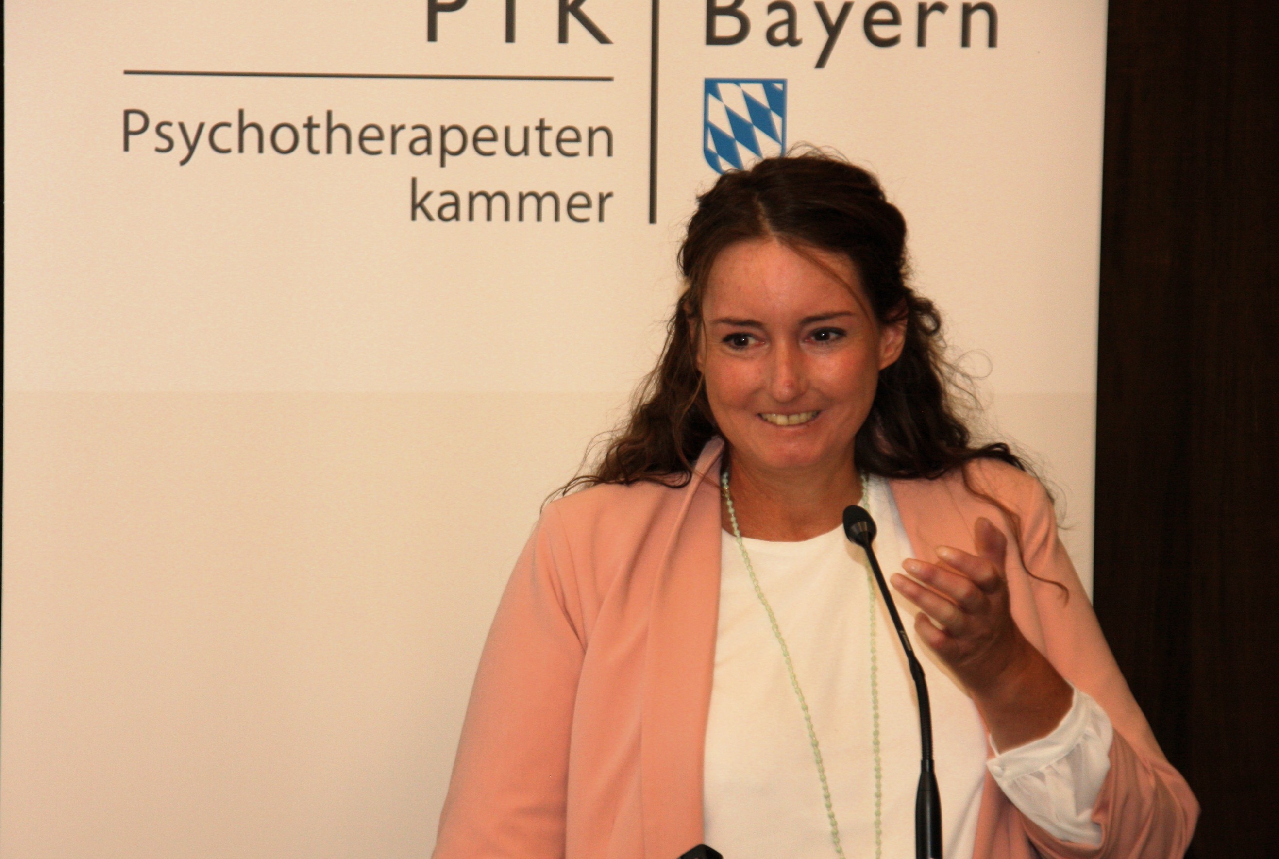 Eine dunkelhaarige Frau in rosafarbenem Blazer steht lächelnd am Redepult. Im Hintergund ist ein Banner der PTK Bayern zu sehen.